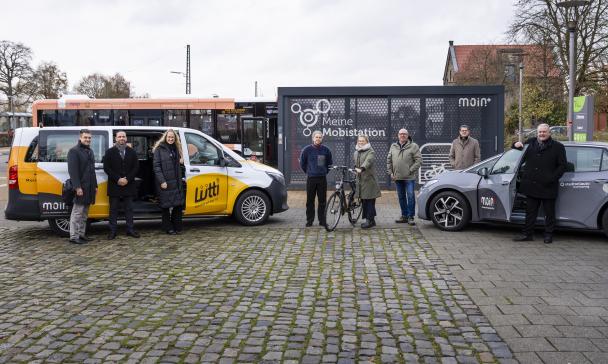 Eine Personengruppe steht vor einem Kleinbus und einem PKW.