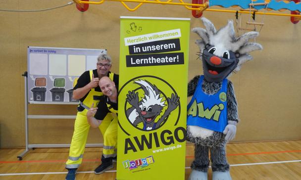 Zwei Personen und ein Maskottchen stehen neben einem Roll Up Poster.