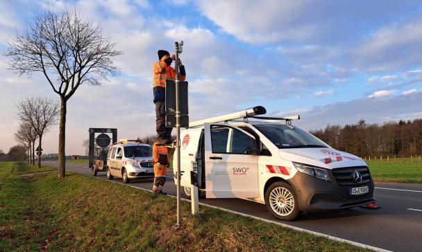 Zwei Kleinbusse parken auf einer Straße. Eine Person montiert einen Sensor an einem Mast.