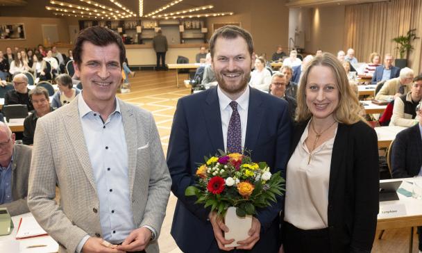 Drei Personen stehen in einem Sitzungssaal.