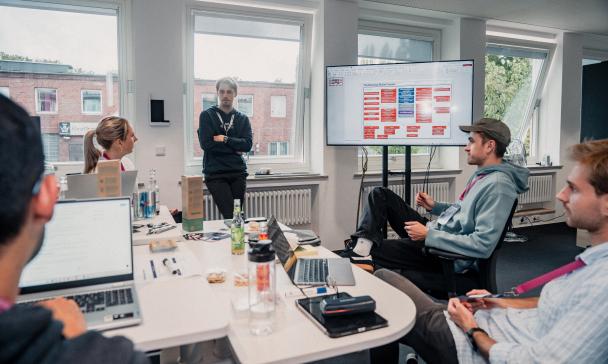 Vier Personen sitzen um einen Besprechungstisch herum, eine steht.