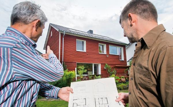 Zwei Personen stehen vor einem haus und halten einen Bauplan in der Hand.